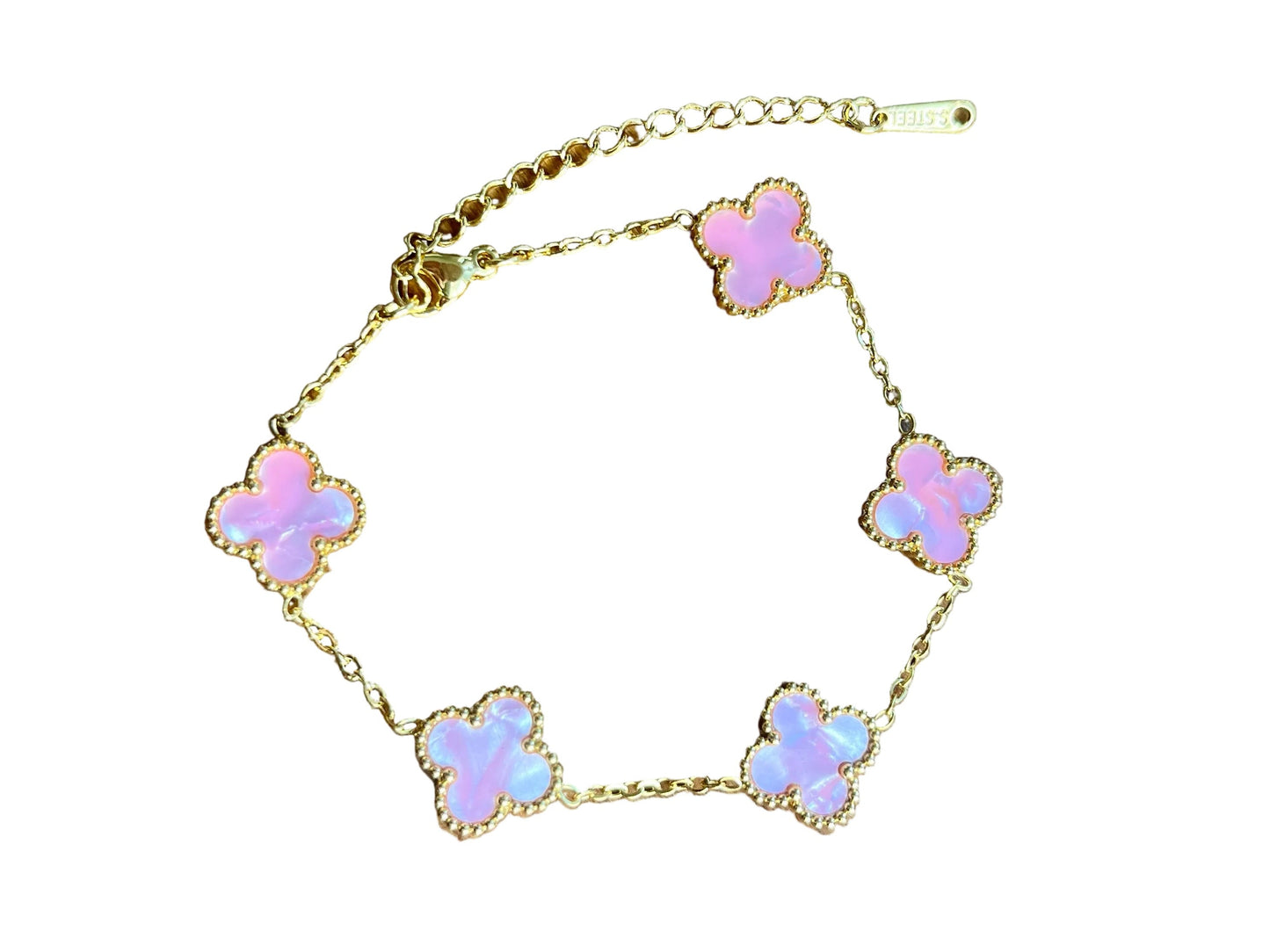 A gold chain bracelet with five small clover-shaped charms in iridescent pink and white, displayed on a wooden surface.